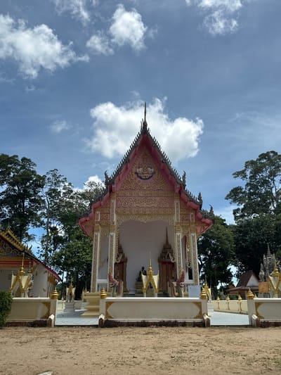 The image depicts a portion of a Buddhist temple, likely in Baan Tha Po, Thailand. The perspective is from ground level looking upwards, with the camera rotated such that the temple's ornate roof dominates the upper portion of the frame. The temple features intricate detailing in pink and gold, with decorative carvings and what appear to be mythological creature motifs along the roofline. White pillars with gold accents support the structure.

In the background, lush green trees provide a natural contrast to the man-made architecture, and a bright blue sky with fluffy white clouds suggests a sunny day. The ground in the foreground is a sandy, earthy color, characteristic of outdoor temple grounds in Southeast Asia. There are no people visible in the frame, suggesting a quiet, perhaps devotional atmosphere.

The overall impression is one of a serene and culturally rich setting. The elaborate decorations and vibrant colors point to a well-maintained and possibly active place of worship. The time of day appears to be daytime, given the bright sunlight and clear sky. No visible text is discernable in the image.
