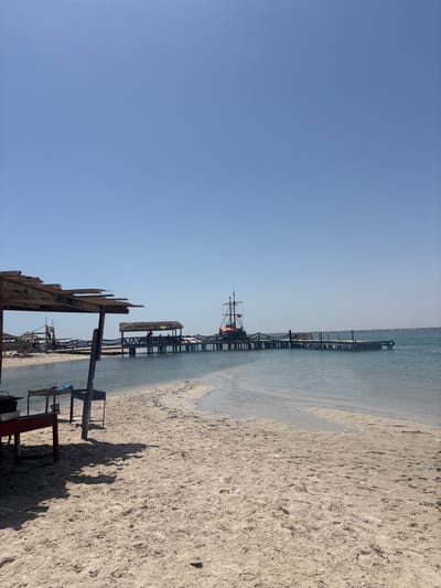 The image depicts a sandy beach with a pier extending into the clear, calm sea. On the left side of the frame, a rustic wooden shelter with a thatched roof provides shade, and beneath it, two red-legged grills are visible. The pier, constructed with wooden planks and railings, supports thatched-roof structures and leads to a docked pirate ship with multiple masts and red flags. Several figures can be seen on the pier, suggesting a recreational area. The sky is clear and blue, indicating fair weather. The scene is characteristic of a coastal establishment in Houmt Souk, Tunisia, likely a restaurant or tourist attraction.