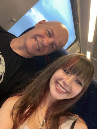 The image is a selfie taken inside what appears to be a train carriage. Two individuals are visible: an older bald man with a black t-shirt and a younger woman with long brown hair, wearing a white top and necklaces. They are both smiling. The background shows a window with a view of the sky and the interior of the train, including lights and seats. There is no indication that this location is in Azor, Israel.