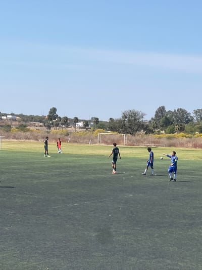The image displays a soccer field in La Venta, Mexico, under a clear, bright blue sky. Six male individuals are visible on a dark green artificial turf surface. Three individuals wear dark green jerseys and shorts; one of these, facing away, has the number "11" on his back. A fourth individual, in an orange jersey and dark shorts, stands further back on a lighter green natural grass area. The remaining two individuals wear blue jerseys with white accents and white shorts; one of them, facing left, holds a white soccer ball in his raised left hand. Two white metal soccer goalposts are present in the midground, situated on the natural grass section of the field. The background features a hilly, arid landscape with sparse vegetation, including trees, shrubs, and occasional residential structures. The scene is illuminated by strong sunlight, casting distinct shadows.