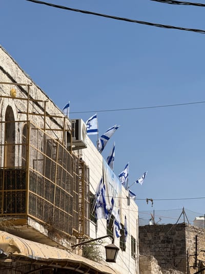 This image presents a daytime outdoor scene in Al-Khalil, Palestinian Territory, under a clear blue sky. To the left, a building constructed of light-colored, rough-hewn stone features an arched window opening and a significant portion covered by a yellowish-brown metal security grate. A white air conditioning unit is mounted on its facade. Below this, a worn, light-colored awning, possibly corrugated metal or fabric, extends outwards. To the right, a multi-story building with a smooth, light-colored facade is prominently adorned with numerous Israeli flags, identifiable by their white field and blue Star of David, displayed along its roofline and upper stories. A large white banner with blue Hebrew text, reading "שבת חברון" (Shabbat Hebron), is partially visible on this facade. A black, ornate street lamp with a lantern-style fixture is affixed to this building. In the background, to the far right, a lower stone wall structure topped with a metal fence and barbed wire is visible, with a white cylindrical water tank positioned behind it. Multiple black electrical power lines traverse the upper section of the image horizontally. No individuals are present within the frame.