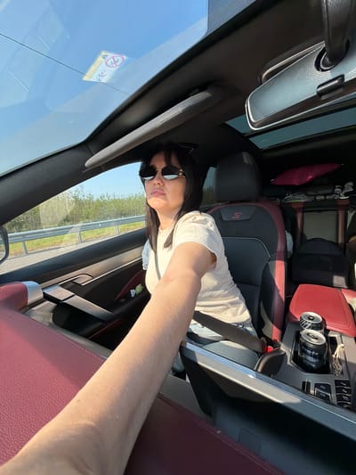 The image shows the interior of a car. A woman wearing sunglasses and a white t-shirt is in the driver's seat. She has dark hair and is looking towards the right. The car has red and black leather seats, and there are two cans of soda in a cup holder. The car is on a road with trees and a guardrail visible outside the window. The sky is clear and blue. The image was taken in Fyodorovo, Russia.