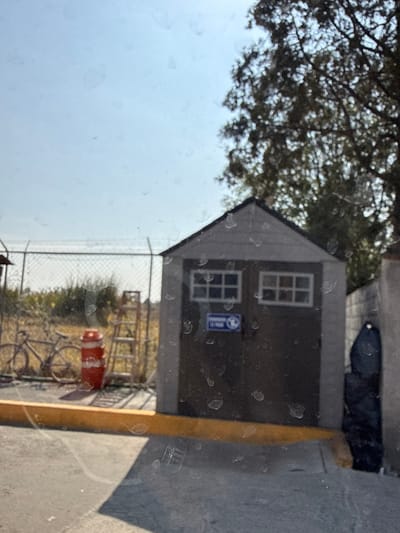 A small building, possibly a shed, has a gabled roof and two windows. A blue sign is placed between the windows. Outside, a chain-link fence runs along the side, partially obscuring a bicycle parked next to a red container. Beyond the fence, vegetation is visible. A tree's branches overhang the building, and a concrete surface runs along the side of the building. The image appears to be taken through a dirty or smudged window.