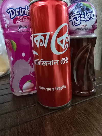 Three beverages are displayed on a wooden surface. From left to right, they are a pink bottle with "Drink Litchi Fruit Drink float With Nata Jelly" written on it, a red can with "Coca-Cola" in white lettering and Bengali text, and a clear bottle containing a dark reddish-brown liquid with a purple label depicting blueberries and Bengali text. The wooden surface appears to be a table. The lighting suggests an indoor environment. The image does not provide any information about the specific location within Chor Sahapur, Bangladesh.