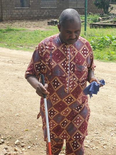 The image shows a man standing outdoors in what appears to be Kabwir, Nigeria. He is wearing a maroon and gold patterned shirt. In his right hand, he holds a blue and white checkered cloth. His left hand is holding a white cane with a black handle, a common tool used by visually impaired individuals.

The man is positioned in front of a brick building on the left side of the frame, with green grass and a sandy path stretching out before him. The background suggests a natural, possibly rural, setting. The lighting indicates it is daytime, likely a sunny day given the clear visibility and shadows.

The man's expression is neutral, and there are no visible text elements in the image. The overall scene depicts an everyday moment for the man, showcasing his use of a mobility aid.