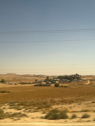 The image depicts a hazy, arid landscape in what appears to be a rural or semi-rural area, likely during daylight hours. The scene is dominated by dry, golden-brown hills and fields, interspersed with sparse green vegetation, suggesting a hot and dry climate. A residential area, with numerous houses and some trees, is visible nestled into the side of a hill. A dirt road winds through the terrain, indicating some level of human settlement and activity.
In the sky, several power lines stretch horizontally across the frame, suggesting the presence of infrastructure. High in the sky, two small, reddish-orange objects are visible; their nature is unclear from the image alone but they could be balloons or possibly drones. The overall atmosphere is subdued due to the haze, which softens the light and reduces visibility, creating a somewhat muted color palette. The location context of al-Qasoum Regional Council, Israel, aligns with the dry, hilly terrain and vegetation observed. There is no visible text in the image.