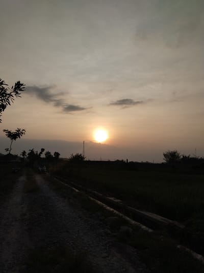 The image shows a rural scene at sunset in Menjing, Indonesia. A dirt road leads into the distance, with dense vegetation on either side. A drainage ditch runs along the right side of the road. Several motorcycles are parked near a small building on the left. In the background, trees and a cell phone tower are silhouetted against the setting sun, which is partially obscured by clouds. The sky is a pale orange color.