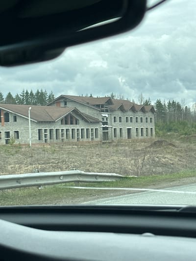 The image, taken from inside a vehicle, captures a large, multi-story building appearing to be either unfinished or abandoned. The perspective suggests it was photographed while traveling past it.

The primary subject is the imposing building, constructed with grey concrete blocks and featuring numerous empty window openings, indicating a lack of glazing or interior finishing. It has multiple brown-tiled pitched roofs with various gables and dormer-like structures, suggesting an architectural style common for large residential or hotel-type properties. The roof appears mostly complete. Surrounding the building, and in the background, are sparse trees, many with bare branches, suggesting either late autumn or early spring foliage. The ground around the structure is covered in dry, light brown grass and some patches of green.

In the foreground, to the right of the building, runs a road or highway, partially visible, with a metal guardrail alongside it. The immediate foreground on the left shows parts of the vehicle's interior, such as the A-pillar and door frame.

The sky is overcast with a uniform layer of light grey clouds, indicating a daytime setting, possibly a cool or mild day. There is no discernible activity taking place at the building itself, reinforcing the impression of stalled construction or abandonment. No people are visible, nor is there any visible text in the image. The scene is located in Bolgury, Russia.