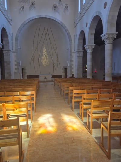 The image depicts the interior of the Church of the Resurrection in Abu Gosh, Israel. The perspective is from the aisle, looking towards the altar. Rows of simple wooden chairs with light-colored seat cushions fill the nave, arranged in parallel lines on either side of a polished, light-colored tiled floor. Sunlight streams through stained glass windows, casting colorful patterns of yellow and orange light on the floor and the fronts of the chairs in the foreground.
The church architecture features large, arched openings supported by stone columns with ornate capitals. The walls are painted a light color, and the ceiling is visible with structural elements. Towards the rear of the church, the altar area is visible with a white cloth-covered altar, a cross, and a vase of flowers. Above the altar, a large arched recess contains a golden mosaic artwork resembling flames or a stylized tree, with thin, dark lines extending upwards. Faint, frescoed figures of angelic or saintly beings are painted on the curved upper walls near the ceiling. The overall atmosphere is one of quiet solemnity and spiritual serenity, enhanced by the interplay of light and shadow within the architectural space.