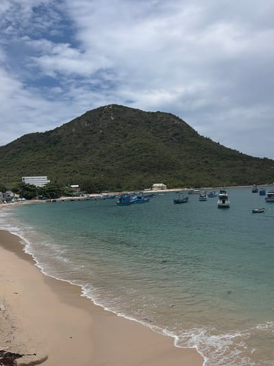 The image depicts a serene coastal landscape in Đông Ninh Hòa, Vietnam. On the left, a majestic, steep mountain covered in lush green foliage dominates the scene, extending its base down to the water's edge. Above the mountain, a bright sky with scattered white and grey clouds is visible.
In the center, a calm body of water, likely a bay or sea, stretches across the frame, displaying various shades of green and blue. Numerous small to medium-sized boats, appearing to be fishing vessels, are anchored or docked in the water, particularly along the base of the mountain. Several modest buildings and structures are discernible along the narrow strip of land between the mountain and the water, suggesting a small coastal settlement or harbor.
To the right, a sandy beach curves along the shoreline, meeting the gentle waves that break softly, forming foamy white lines. The sand appears light brown. No people are clearly visible, and the boats appear stationary, suggesting a tranquil moment in a working coastal environment. There is no discernible text in the image. The scene captures the peaceful coexistence of nature and human activity in a coastal Vietnamese setting.