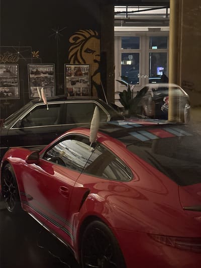 A red Porsche, parked indoors, is the main subject of the photograph. The car's exterior is prominently featured, with the front fender, wheel, and headlight assembly being clearly visible. The vehicle has racing stripes.
In the background, there is a wall with a graphic design featuring a lion's head and a collage of vintage photographs. To the left, a window shows an external night scene.
Based on the presence of a red Porsche, wall graphics, and architectural features, the location is likely within the "Werk 1" building in Wetzikon (ZH), Switzerland. This building is known for housing a car workshop and vintage car storage.