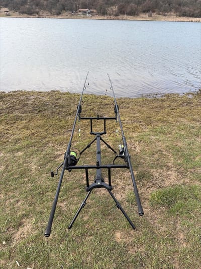 The scene is a lakeside fishing setup in Cherna Gora, Bulgaria. In the immediate foreground, a black, three-legged fishing rod pod supports two dark fishing rods, positioned horizontally and angled towards the water. Both rods are equipped with reels spooled with yellow-green fishing line, and bite indicators featuring yellow-green elements are suspended from the lines. The ground beneath the equipment consists of sparse, dry grass and patches of exposed earth. The midground features a calm body of water with minimal surface ripples. On the distant bank, a line of bare deciduous trees stretches horizontally, partially obscuring a small, light-colored building with a dark roof, likely a hut, visible towards the left. No individuals are visible within the frame.