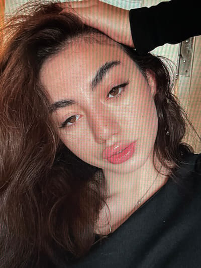 The image is a close-up, head-and-shoulders shot of a young woman indoors, with her right hand resting on top of her head. She has long, dark brown hair that falls over her left shoulder and across her face. Her eyes are brown and she is looking towards the camera. She is wearing makeup, including eyeliner and eyeshadow, and her lips are glossy and pink. A silver necklace is visible around her neck. She is wearing a black top. The background is indistinct and appears to be a wall with some vertical lines, possibly a grate or part of a window. There are no visible elements to suggest a specific location in Chotěšov, Czechia.