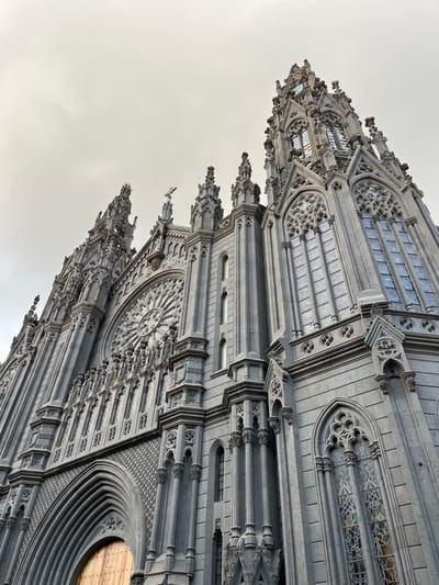 The image shows the exterior of a large, gothic-style church building in Arucas, Spain. The church is constructed of gray stone and features a large rose window, a central spire, and numerous other ornate details. The building is a prominent landmark in the city and is located at 28.119 latitude, -15.523 longitude. It appears to be a cloudy day. The church is an architectural marvel and is a testament to the skill of the builders. The church is the Iglesia de San Juan Bautista, which is one of the most important churches in Arucas. The church is a popular tourist destination, and visitors can admire its intricate architecture and learn about its history.