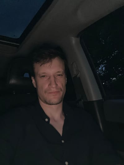 This is a slightly tilted, low-light, close-up indoor photograph of a young man. He is wearing a black collared shirt with two buttons visible. His hair is dark and appears somewhat messy. He has stubble on his face and a light tan. His expression is relaxed and he is looking directly at the camera with a slight smile.
The background suggests he is inside a vehicle. Through a window on the left, a dark, cloudy sky is visible, indicating it is likely nighttime or very late evening. There are also reflections on the window, possibly from raindrops, suggesting it may be raining or has recently rained. The overall lighting is dim, with the man's face being the primary illuminated subject. The setting appears to be Makiv, Ukraine. No text is visible in the image.