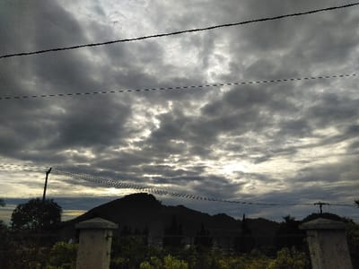 The image displays a mountainous landscape in Hafjell, Norway, under an overcast sky. The upper two-thirds of the frame consist of dense, dark grey stratocumulus clouds, with several patches of brighter sky and sunlight visible through gaps, particularly towards the horizon. Two prominent black wires, consistent with electrical or telecommunication lines, traverse horizontally across the upper portion of the sky, with a fainter dashed line below the topmost wire.

In the immediate foreground, two weathered, light-colored concrete fence posts are partially visible on the left and right sides of the frame, suggesting a barrier or gate structure. Below and between these posts, there is dense, dark green foliage and vegetation. A dark utility pole, likely wooden, stands on the left behind the foreground vegetation, supporting multiple horizontal lines.

The midground features the distinct, dark silhouette of a large mountain peak situated to the center-left, with its crest defined against the lighter sky. To its right, additional undulating hills and mountains extend across the horizon, also appearing as dark masses. Scattered dark coniferous trees are discernible in the lower midground, in front of the mountains. No people, specific actions, or interactions are observed within the scene.