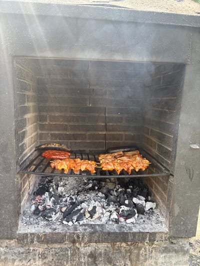 An outdoor brick barbecue pit is filled with smoldering coals. A metal grill rack is positioned above the coals, holding various items being cooked. On the left side of the rack, there appear to be two long sausages, possibly chorizo, and a longer, darker item that could be a vegetable like an eggplant or plantain. To the right of the sausages, several skewers of marinated chicken pieces are arranged. A thin wisp of smoke rises from the barbecue. The barbecue pit itself is constructed from dark brick and has a stone or concrete facade. The background is out of focus but suggests an outdoor setting. The image is taken from a slightly elevated perspective, looking down into the barbecue.