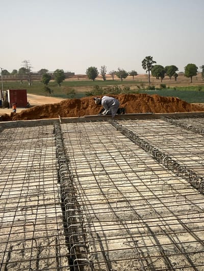 The image, when viewed with the correct orientation (rotated 90 degrees counter-clockwise), shows an outdoor construction scene on a bright, sunny day. The dominant feature in the foreground and extending across the right half of the frame is a dense, intricate grid of steel reinforcement bars (rebar). This rebar framework is laid out flat, suggesting preparation for pouring a concrete slab or a foundation. The grid consists of intersecting horizontal and vertical bars, with sections featuring additional rows of U-shaped or rectangular stirrups, indicating areas of higher structural reinforcement, possibly for beams or columns embedded within the slab. The surface beneath the rebar appears to be a pre-existing concrete layer or formwork.
Adjacent to the rebar structure, running along the left edge of the main construction area, is a strip of reddish-brown soil or dirt, forming an embankment or boundary. On this dirt strip, in the midground, a worker is seen kneeling. The worker is dressed in light-colored clothing, possibly a white or light grey long-sleeved shirt and trousers, and is facing towards the rebar, seemingly engaged in a task related to its installation, inspection, or tying.
The background opens up to a wide, flat landscape under a clear, light-colored sky. Scattered across this open area are numerous trees of various sizes and shapes. A tall tree with a distinct fan-like crown, resembling a palm tree, stands out in the left-mid background. Other trees, mostly green-leafed, are interspersed across the landscape, which features patches of dry, light-colored earth and some areas of green vegetation, possibly fields or natural growth. In the far left background, near the horizon, a red object that resembles a shipping container or the back of a truck is visible, with a small human figure standing nearby. Faint structures or buildings can be discerned in the distant background.
The scene conveys a sense of active construction work taking place in a relatively open, possibly rural or semi-urban environment, under clear, bright daylight conditions. There is no visible text in the image.