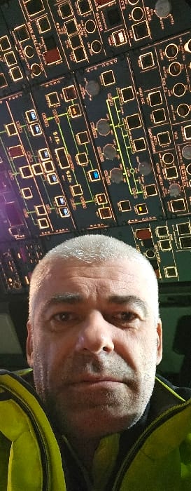 A male individual is depicted in an aircraft cockpit at Katowice Airport (KTW) in Pyrzowice, Poland. The subject, a middle-aged to older male with short, light grey hair, is visible from the chest up, positioned in the lower half of the image. He wears a high-visibility lime-green or yellow jacket over a darker garment. His facial expression is neutral, and he is looking directly into the camera, indicative of a self-portrait. Above and behind the individual's head, an illuminated overhead control panel of an aircraft is prominently displayed. The panel is dark, featuring numerous square and circular buttons, switches, and interconnected diagrams, all emitting an amber glow. Several indicators on the panel are also illuminated with blue light.