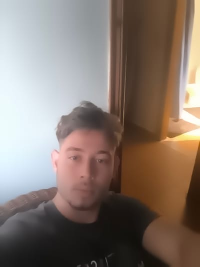 The image shows a medium close-up of a young man with brown hair and eyes, wearing a dark t-shirt. He is sitting in a chair, with a light blue wall behind him and a doorway with brown trim to his right. The lighting suggests it is daytime, with light coming from the right side of the frame. The man appears to be looking at the camera with a neutral expression. Partially visible on his left shoulder is the head and neck of a snake, with patterned scales. The background is blurred, with a bright yellow light source visible in the distance on the right. There is no visible text. The image quality is somewhat blurry, particularly around the edges.