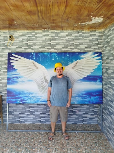 A man wearing a yellow bucket hat, a blue t-shirt, and tan shorts stands in front of a large mural of white angel wings against a blue, cloudy sky and water background. The man has a beard and a slight smile. He is wearing sandals. The mural is propped up by a metal frame. Behind the mural is a gray brick wall. Above the brick wall is a ceiling that appears to be made of wood planks with some damage or discoloration. The floor is tiled with a mosaic of brown, tan, and gray stones. The scene appears to be outdoors or in an open-air structure, possibly in K'Rèn, Vietnam, given the context.