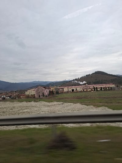 The image captures an industrial complex in Cherniche, Bulgaria, viewed from a moving vehicle under an overcast sky. In the foreground, a gray metallic guardrail runs horizontally, with blurred green grass visible beneath it. Beyond the guardrail, an expanse of light-colored, disturbed earth leads to the industrial area. On the left, a multi-story building with a faded reddish-pink facade and rectangular windows stands. Adjacent to it are stacked materials and several parked vehicles. To its right, a series of elongated, light-colored industrial buildings with red roofs stretches across the midground. Multiple chimneys or vents on these structures are actively emitting white smoke or steam. The background features rolling, tree-covered hills, with a distinct peak on the far right exhibiting a small structure. Distant, snow-capped mountains are faintly visible beneath a uniformly gray, cloudy sky. No human subjects are discernible. Reflections, consistent with photography through a vehicle window, are present in the upper portion of the frame.