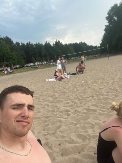 The image depicts a sunny day at a sandy beach, possibly near a wooded area, given the trees visible in the background. Several people are present, engaged in various activities. In the foreground, a person is taking a selfie, looking towards the camera with a relaxed expression. Further in the background, a group of people are gathered on the sand. One person is sitting down, while others are standing or lying down, suggesting a casual social gathering. A volleyball net is set up, hinting at the possibility of recreational games. The weather appears to be fair and bright, typical of a summer day. The overall scene conveys a sense of leisure and enjoyment in an outdoor, natural setting. There is no visible text in the image.