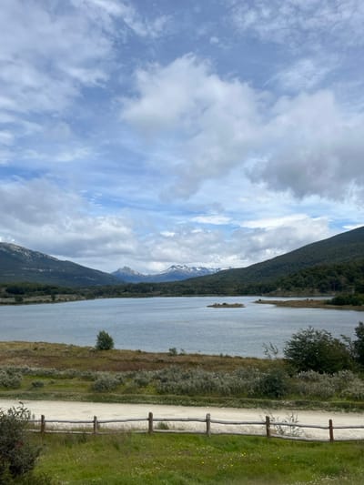 A landscape photo displays a lake next to mountains under a cloudy sky. A walking path runs along the lake's edge, bordered by a rustic wooden fence on one side and natural vegetation on the other. Distant snow-capped mountain peaks are visible. The sky is filled with a mix of white and gray clouds, with patches of blue peeking through. The scene evokes a sense of tranquility and outdoor adventure.
