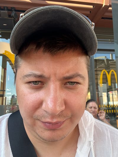 A close-up, selfie-style photograph shows a fair-skinned man wearing a dark grey baseball cap with sunglasses perched on top. He has short, light brown hair visible beneath the cap and light stubble on his upper lip and chin. His eyes, with green irises, are looking directly at the camera. He is wearing a white shirt, and a black strap is visible over his left shoulder. In the background, there is a glass building with a yellow McDonald's logo visible. Behind the man, a blurred image of an older woman with dark hair and a patterned top is also visible. The overall setting appears to be outdoors, with a structure of wooden slats overhead. The presence of the McDonald's logo suggests a location in or near Sedaví, Spain.
