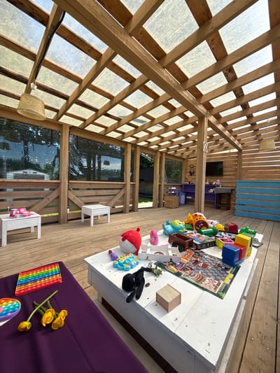The image shows an outdoor wooden gazebo with a translucent roof, located in the city of Zamosh'e (Zaklinskoe S/P), Russia. The gazebo is furnished with wooden decks, tables, and seating. Several children's toys are scattered on the tables and the floor, including building blocks, toy cars, a stuffed animal, and pop-it toys. A purple cushion with yellow flowers is visible in the foreground. In the background, there are shelves with additional toys and a television screen mounted on a wooden wall. The overall impression is a playful and casual outdoor space, possibly a play area or a backyard recreational zone.