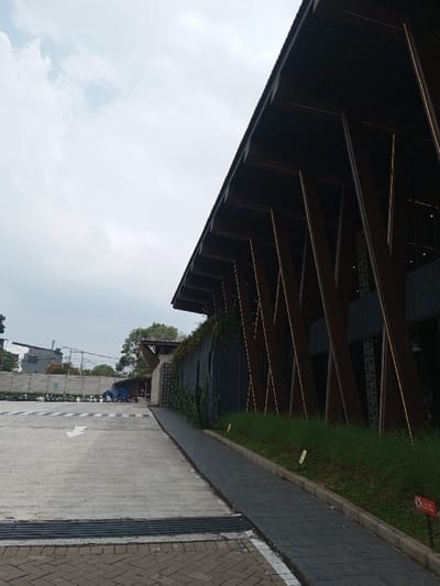 This image depicts an outdoor architectural scene within Tân Kỳ Commune, Vietnam. The right side of the frame is dominated by a modern building featuring a dark, steeply sloped roof and an angular facade composed of numerous dark vertical support beams, each exhibiting a thin, integrated horizontal light strip. Sections of the building's dark gray wall are partially covered by green foliage. Adjacent to the building, a strip of green grass contains thin, tall vegetation and small yellow ground lights, bordering a dark gray paved pedestrian pathway. A partially visible red sign with a white pictogram is situated at the bottom right. To the left, a wide expanse of light gray concrete, marked with a white forward-pointing arrow and a distant pedestrian crossing, extends towards the background. A dark, segmented grate spans the transition between the concrete and a foreground section of dark gray brick pavers. The background reveals a parking area containing multiple parked motorbikes, low concrete walls, and distant urban structures and trees under an overcast sky. No human figures or discernible actions are present.