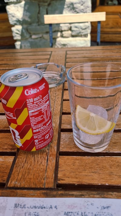A close-up shot shows a Coca-Cola can and a glass of ice with a lemon slice on a wooden table. The Coca-Cola can is red and yellow, with "Coke" written in white on the top. The glass is clear and contains several ice cubes and a thin slice of lemon. An ashtray is visible behind the Coca-Cola can. The table appears to be outdoors, possibly on a patio, with a stone wall and a wooden bench visible in the blurred background. The scene suggests a casual setting, likely in Rupit, Spain, given the visible text on the can which includes Spanish language. The lighting indicates it is daytime.