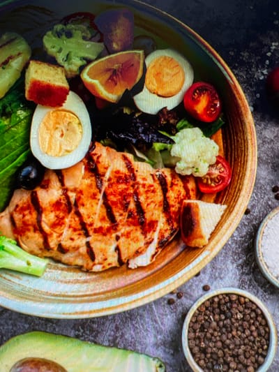 The image depicts a close-up shot of a salad bowl, with a focus on the food composition. The bowl contains grilled chicken breast with visible grill marks, halved hard-boiled eggs, sliced cherry tomatoes, small florets of cauliflower, mixed greens (including darker leafy varieties), avocado pieces, and what appears to be a slice of toasted bread or crouton. The ingredients are arranged within a shallow, wide-rimmed bowl with a textured, rustic appearance. To the side of the bowl, there are small bowls containing peppercorns and salt crystals, along with half an avocado. The background consists of a textured, dark surface. There is no indication of this image being located in Blackburn, United Kingdom. No people, actions or interactions are evident.