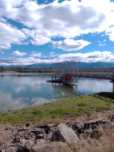 The image shows a landscape scene in Municipio Tarija, Bolivia. The foreground is characterized by a rocky, uneven terrain, covered in grass and vegetation. Further ahead, a body of water, possibly a lake or reservoir, stretches towards the middle ground. Reflections of the sky and clouds are visible on the water surface. An elevated structure or bridge extends into the water from the shoreline. In the background, mountains form the horizon line, with a clear sky and scattered clouds above.