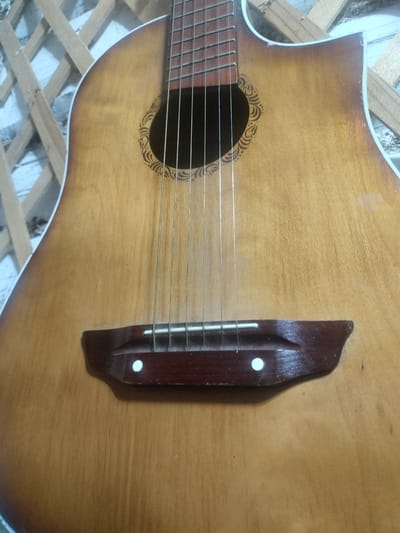 The image shows a close-up of an acoustic guitar. The guitar has a light brown wooden body with a darker brown gradient towards the edges. The soundhole is round and has a decorative pattern around it. Six strings are visible, running from the bridge to the headstock. The bridge is made of dark wood and has white dots embedded in it. The background consists of a light-colored wall with a wooden lattice structure. The location in Akbasheva, Russia, is not discernible from the image itself.