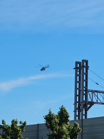 A small, light-colored helicopter is seen in flight against a clear blue sky in the city of Trekhgorka, Russia. The helicopter is positioned in the upper left quadrant of the image. To the right of the helicopter, a tall, metallic structure, possibly part of a power line pylon or bridge support, rises from the lower right corner. The pylon is grey and has a hollow, rectangular design. In the foreground, partially obscuring the base of the pylon and extending across the bottom of the image, is a grey, corrugated barrier. Two green, leafy trees are visible in the lower left and lower right corners, with the tree on the left being more prominent. A faint white contrail or cloud is visible in the sky behind and to the left of the helicopter. The overall scene depicts an open, outdoor environment under daylight conditions.