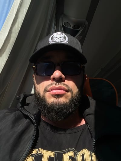 A man with a beard and sunglasses is sitting inside a vehicle. He is wearing a black baseball cap with a white skull logo, a black zip-up jacket over a black t-shirt with a yellow graphic. The vehicle appears to be a bus or train, with seats and windows visible. Sunlight streams through the window, casting shadows on the man and the interior of the vehicle. The man is looking directly at the camera, and his facial expression is neutral. The location is the city of Vitino, Russia.