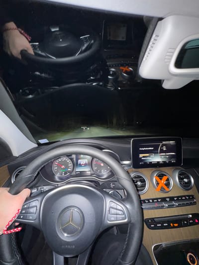 The image depicts the interior of a Mercedes-Benz vehicle from the driver's perspective at night, located in Bohot, Bulgaria. A left hand, adorned with a red and white braided bracelet, grasps the black leather-wrapped steering wheel. The instrument cluster shows the vehicle traveling at 63 km/h, with the tachometer indicating approximately 1200 RPM. The central infotainment screen displays media information, specifically "Facing RediculousOfficial" and "Anton's iPhone". The dashboard features a wooden trim and three circular air vents, one of which is marked with an orange "X" sticker. An identical orange "X" is visible as a reflection on the windshield in the upper right quadrant. Outside the vehicle, headlights illuminate a dark, unpaved road extending into the distance, flanked by trees or dense vegetation under a clear night sky. A faint, small illuminated object or marker is visible ahead on the road. Reflections of the dashboard and a partial reflection of the driver's hand are visible on the windshield.