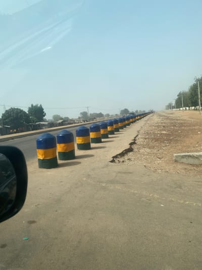 The scene depicts a straight, two-lane asphalt road in Yan Dala, Nigeria, observed from within a vehicle, with a portion of a side mirror visible in the lower left foreground. The sky above is a hazy, light blue. Along the right edge of the paved road, a continuous line of approximately 15-20 cylindrical concrete bollards extends into the distance. Each bollard is painted in three horizontal bands: blue on top, yellow in the middle, and green at the base.

Adjacent to the bollards, the unpaved roadside consists of dry, reddish-brown dirt and gravel, exhibiting noticeable erosion and a fissure along the edge of the asphalt. Further right, utility poles and scattered, slender trees line the path, along with some concrete fragments on the ground. To the left of the paved road, across the thoroughfare, are various low-rise structures, possibly commercial or residential, interspersed with green vegetation. The distant background on both sides of the road is obscured by atmospheric haze, revealing only faint outlines of structures and vehicles. No human figures or discernible actions are present.
