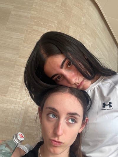 Two females are depicted in a close-up, indoor shot against a light beige tiled wall. The background tiles are rectangular and arranged in a stacked bond pattern. The upper subject, a female with long dark hair parted centrally, is positioned with her head resting on the crown of the lower subject's head. She wears a white long-sleeved athletic shirt with a black Under Armour logo visible on the right chest area. Her gaze is directed towards the viewer, featuring dark brown eyes and visible eyebrows. The lower subject, also a female, has brown hair parted centrally and blue eyes. She wears a dark-colored top and a white pearl necklace. Her expression involves a pouting mouth and her eyes are rolled upwards and to the viewer's left. She holds a green glass bottle with a silver cap that features a red fleur-de-lis emblem, positioned in the lower left portion of the image. The scene is located in the city of Berry Islands, Bahamas.