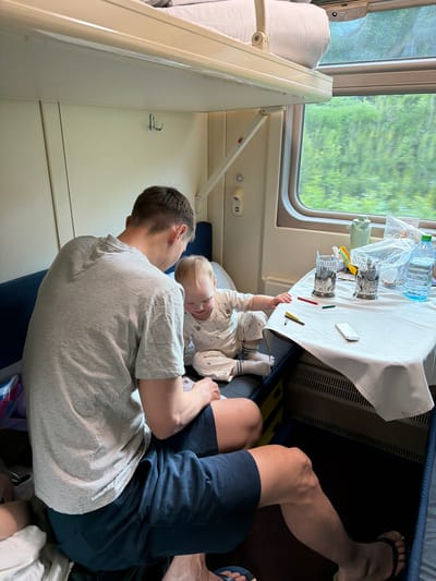 A man is sitting down, wearing a grey t-shirt and navy shorts, with a baby on his lap. The baby is wearing white pajamas with a pattern on them. The man and baby are seated on a blue cushioned surface. Beside them is a small table covered with a white tablecloth. On the table are multiple glasses, a water bottle, and several small items including writing utensils. The image appears to be taken inside a train car, as evidenced by the window showing a green landscape passing by, as well as the presence of a sleeping bunk. The train compartment contains two levels of sleeping bunks.