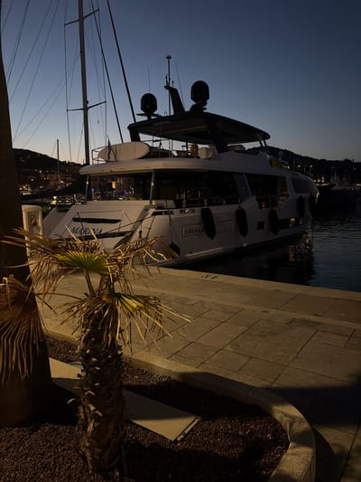 A modern, white yacht named "MARINA" is moored at night in the Port of Imperia, Italy. The yacht, which also has "SIRENA" visible on its hull, is illuminated by internal and external lights, casting reflections on the dark water. Several black fenders are attached to the yacht's side. A paved walkway, likely part of the marina, runs along the edge of the water, with a low concrete border containing dark gravel and two small palm trees. One palm tree in the foreground has dry, brown fronds, while the other is partially visible to its left. In the background, other boats and masts are visible, along with lights from the city lining the hillsides. The sky is a deep twilight blue. The scene depicts a still, quiet evening at the harbor.