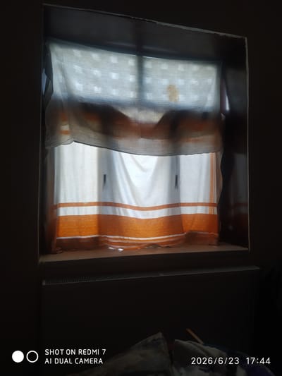 The image shows a window with curtains. The curtains are made of two layers: an outer layer that is sheer and off-white, and an inner layer that is white with orange stripes. The outer layer is pulled to the side, revealing the inner layer. Through the sheer outer curtain, a faint grid pattern is visible, suggesting it is a window looking out onto a building with many windows. A brownish stain is visible on the sheer curtain. The window sill is visible below the curtains, and a radiator is located beneath the window. The scene is dimly lit, suggesting it is either early morning or late evening. The text "SHOT ON REDMI 7 AI DUAL CAMERA" and a date and time stamp "2026/6/23 17:44" are superimposed on the lower part of the image, indicating the photograph was taken with a Redmi 7 phone at the specified date and time. The location context provided is Khalkabad, Uzbekistan.
