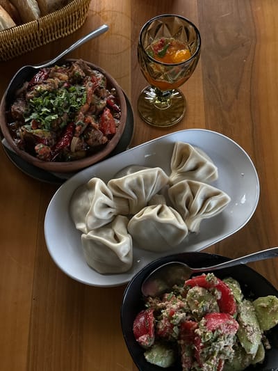 The image shows a wooden table set with a selection of Georgian dishes, likely in the city of Marneuli, Georgia.
On the table are:
* A white oval dish containing six khinkali, which are Georgian dumplings.
* A dark brown ceramic bowl filled with a stew-like dish. The stew contains pieces of meat, tomatoes, and red peppers, garnished with chopped herbs. A spoon is placed next to the bowl.
* A black bowl filled with a salad made from sliced tomatoes and cucumbers, tossed in a creamy sauce and garnished with herbs. A spoon is inside the bowl.
* A glass with a drink that contains sliced oranges.
* A wicker bread basket with slices of white bread.
The dishes and utensils are arranged in a manner typical of a shared meal. The scene suggests a dining experience centered around Georgian cuisine.