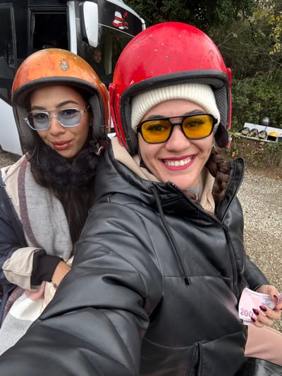 Two women are positioned in the foreground, looking directly at the camera, in an outdoor setting in Çiftlikköy, Turkey. The woman on the left wears an orange full-face helmet, clear-framed sunglasses with blue-tinted lenses, a dark fur-like collar, a striped scarf (white, pink, and beige), and a dark-colored jacket. She has dark hair and a slight smile. Behind her, a white vehicle with dark windows and a red and white logo is partially visible, with a male figure looking out from within. The woman on the right wears a red full-face helmet over a white knit beanie, black-framed sunglasses with yellow-tinted lenses, a black puffer jacket, and a beige hoodie or neck warmer. She displays a broad smile, revealing her teeth, and has dark hair in a visible braid on her right side. Her left hand, adorned with dark red, glossy, almond-shaped fingernails, holds two 200 Turkish Lira banknotes. In the background, green foliage is visible, along with several other helmets (black, yellow, white, grey) resting on a low ledge or wall. The ground appears to be a gravel or dirt surface. The scene suggests a recreational activity involving helmets, possibly ATV riding.