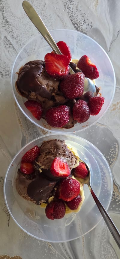 Two clear plastic containers filled with ice cream and strawberries are arranged on a patterned surface. The container on the left holds scoops of what appears to be chocolate and vanilla ice cream, topped with several whole and sliced strawberries. A silver spoon is partially submerged in the ice cream. The container on the right also contains chocolate and vanilla ice cream with strawberries, and a silver spoon is visible resting on top. The background shows a marbled pattern, suggesting an indoor setting. There is no visible text in the image. The lighting suggests it is daytime.