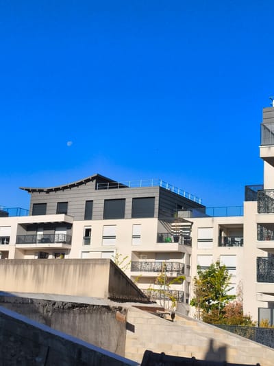 The image depicts a section of a modern residential building complex with a clear blue sky as the backdrop. In the upper left quadrant of the sky, a pale, crescent moon is visible. The building features multiple levels with balconies, characterized by dark metal railings with intricate patterns. The facade is a light, neutral color, possibly beige or cream, with numerous windows, many of which are covered by roller shutters. One building section at the top has a darker, possibly grey, angled roof structure with horizontal detailing.
The foreground shows a contrasting, older structure with a rough, textured beige wall and what appears to be a concrete or stone surface below it. This older structure occupies the lower portion of the image, creating a sense of depth and layering between the foreground and the modern buildings. Green foliage, including trees and smaller bushes, is interspersed throughout the balconies and around the base of the buildings, suggesting a well-maintained environment.
This scene is located in Lagny-sur-Marne, France, specifically in the area of the "Parc des Cèdres," an urban development known for its contemporary architecture.