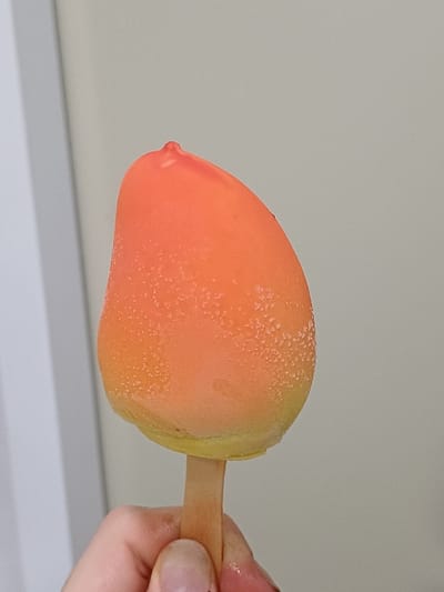 The image presents a close-up, eye-level perspective of a single frozen dessert, identified as a popsicle, held by a human hand. The popsicle exhibits a teardrop or mango-like shape, transitioning in color from a vibrant orange at its apex to a lighter yellow at its base. Its surface is covered with a uniform layer of fine ice crystals and appears slightly wet due to condensation or minimal melting. A light brown, flat wooden stick protrudes from the base of the popsicle. A human hand, with visible knuckles and a light skin tone, grasps the wooden stick with fingers positioned around it. The background consists of a plain, light gray wall. A vertical white architectural element, likely a door frame or wall corner, is partially visible along the left side of the frame. The scene is situated in 서대문구, South Korea.