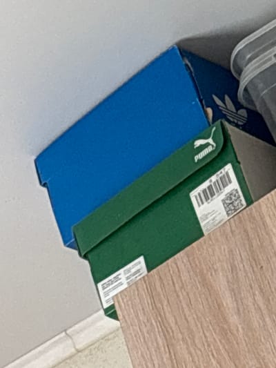 The image displays two shoeboxes, one blue and one green, stacked on a wooden surface. The blue box is on top and partially obscures the green box. The green box has a Puma logo and a label with a barcode and QR code. In the background, a clear plastic container is visible. The scene appears to be indoors, possibly a closet or storage area, with a wall and skirting board in the lower left corner. The image is shot from a slightly elevated angle. There are no people present in the image. The location is not specified in the image.