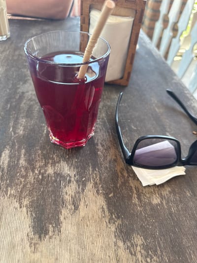 The image shows a close-up of a wooden table outdoors, likely at a cafe or restaurant. On the table is a glass of dark red liquid with a straw, a napkin dispenser with napkins, and a pair of black sunglasses resting on a few napkins. The table surface is weathered and shows visible wood grain and scratches. In the background, there are glimpses of people and what appears to be a white fence or railing, suggesting an outdoor seating area. The context of Puhoi, Moldova, is not explicitly visible in the image itself, but the description aims to be factual based on the visual elements.